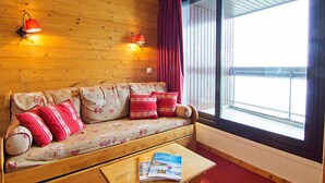 A cozy room with a wooden wall, a sofa with patterned cushions, a small table with magazines, and a large window with curtains.