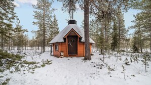 Eine kleine Holzhütte mit schneebedecktem Dach, umgeben von hohen Kiefern in einem verschneiten Wald.