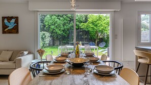A dining area with a wooden table set for six, surrounded by chairs, with a view of a lush garden through large windows. - Les Marines by Interhome (Carnac)