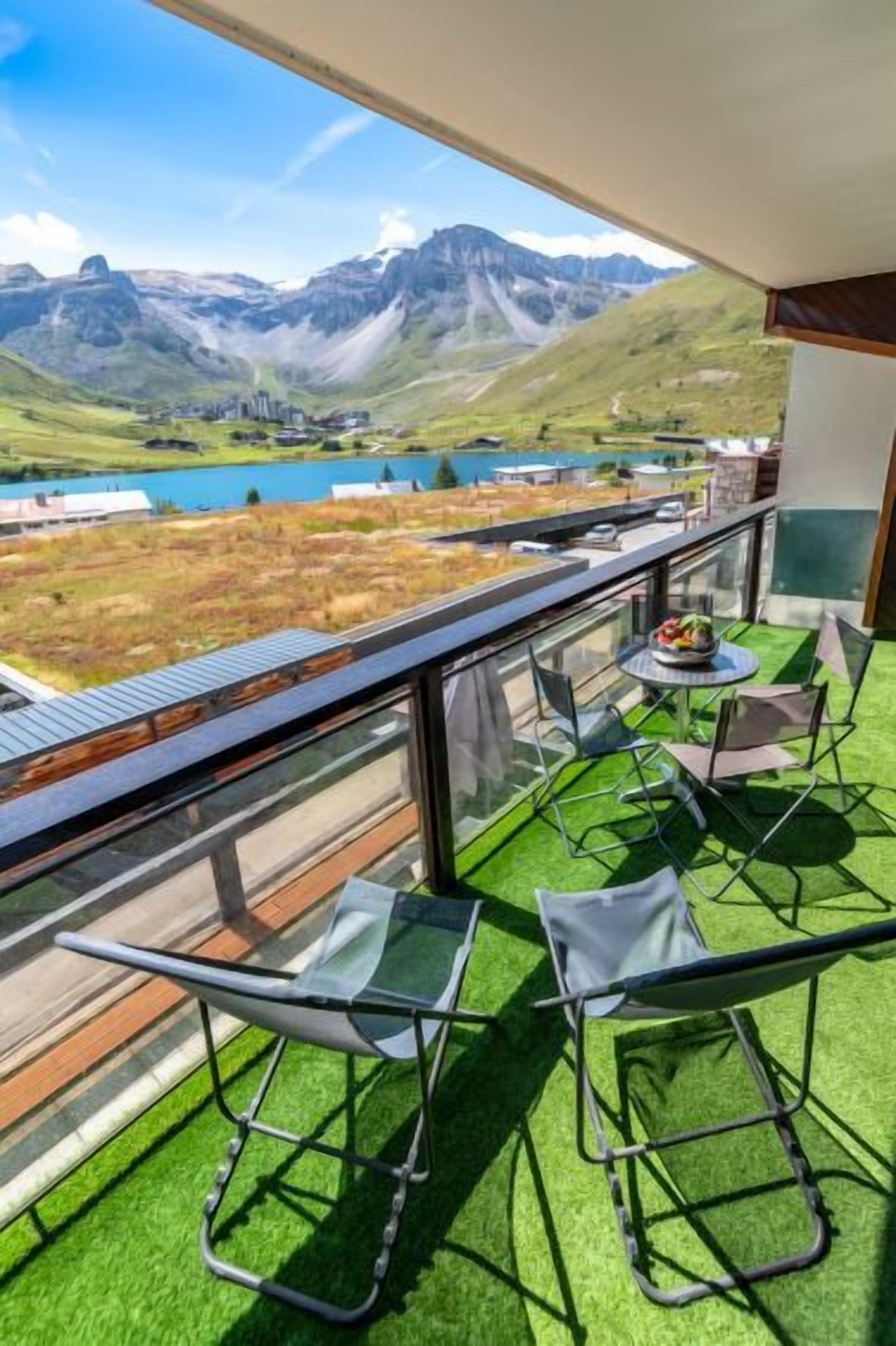 A balcony with a view of a lake and mountains, two chairs, and a table with a bowl of fruit.