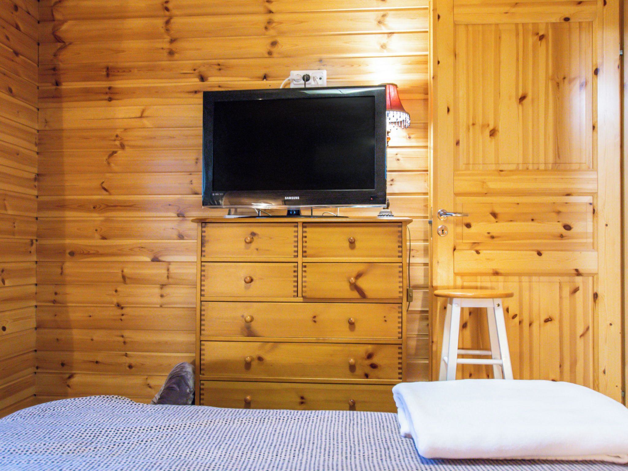 Une pièce aux murs en bois, un lit recouvert d’une couvre-lit à motifs, un téléviseur posé sur une commode en bois et un tabouret blanc.