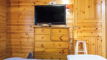 Une pièce aux murs en bois, un lit recouvert d’une couvre-lit à motifs, un téléviseur posé sur une commode en bois et un tabouret blanc.