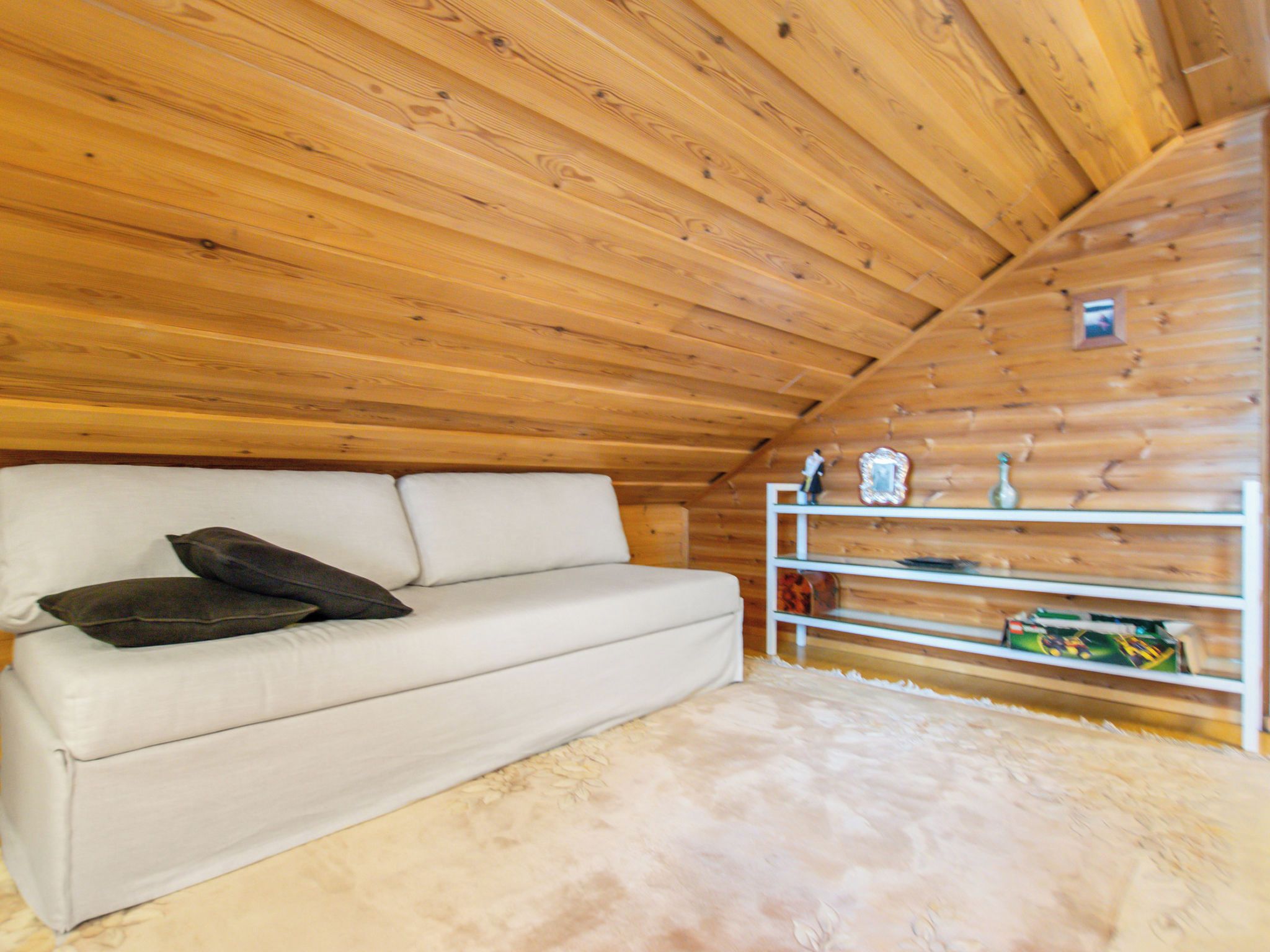 Quarto com sofá branco, tapete felpudo, vigas de madeira no teto e prateleira branca com diversos objetos.