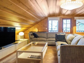 Interior acogedor de una cabaña con techo de madera, un sofá, una mesa de centro, un televisor y una ventana con vista a los árboles.