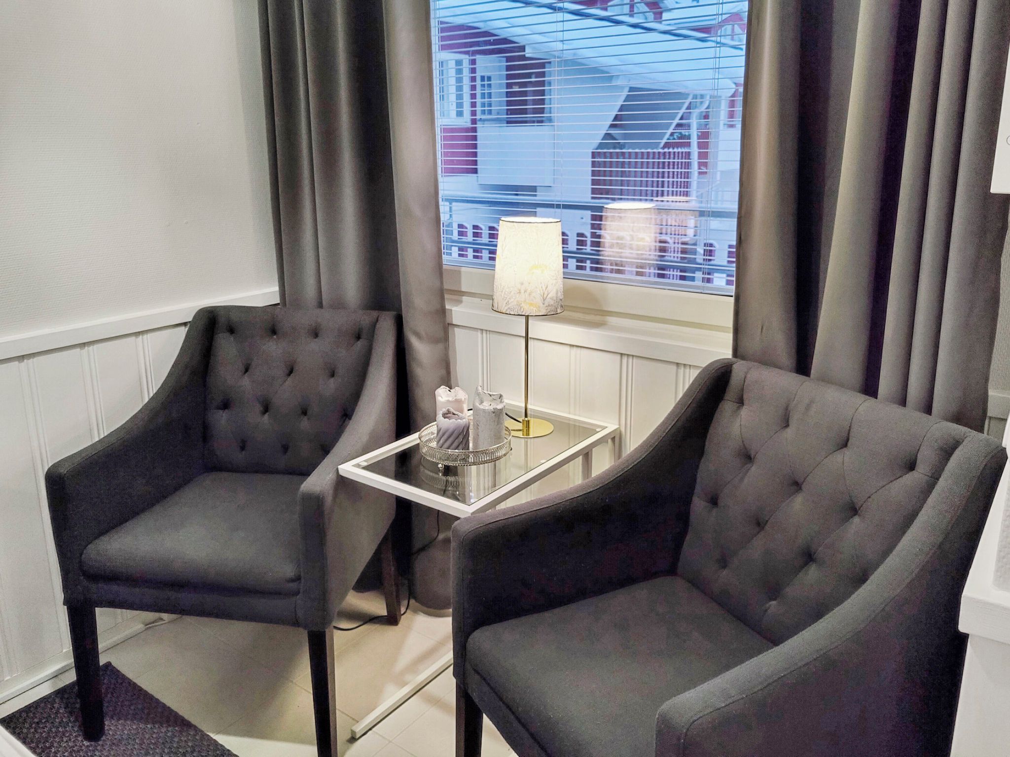 A hotel room with two grey upholstered armchairs, a small glass table with a lamp, and a view of a building through the window.