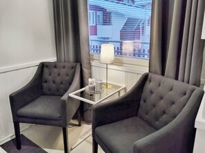 Habitación de hotel con dos sillones grises tapizados, una mesita de vidrio con una lámpara y vistas a un edificio a través del ventanal.