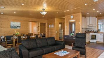 Interior acogedor de una cabaña con sofá, sillas, una mesa de comedor, una zona de cocina y techo de madera.