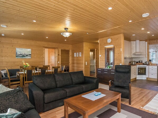 A cozy cabin interior with a sofa, chairs, a dining table, a kitchen area, and a wooden ceiling.