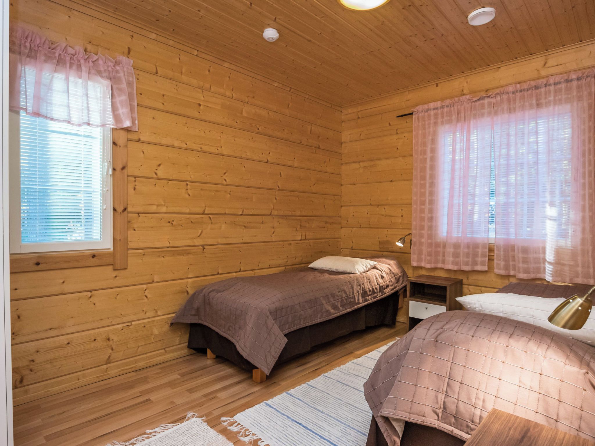 Ein Zimmer einer Holzhütte mit einem Einzelbett, einem Nachttisch, einer Lampe und einem Fenster mit Vorhängen.