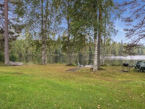 Un parque junto al lago con césped, un banco, un bote y una parrilla.