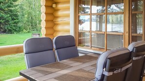 A log cabin with a wooden deck, a table with four chairs, and a large window with blinds.