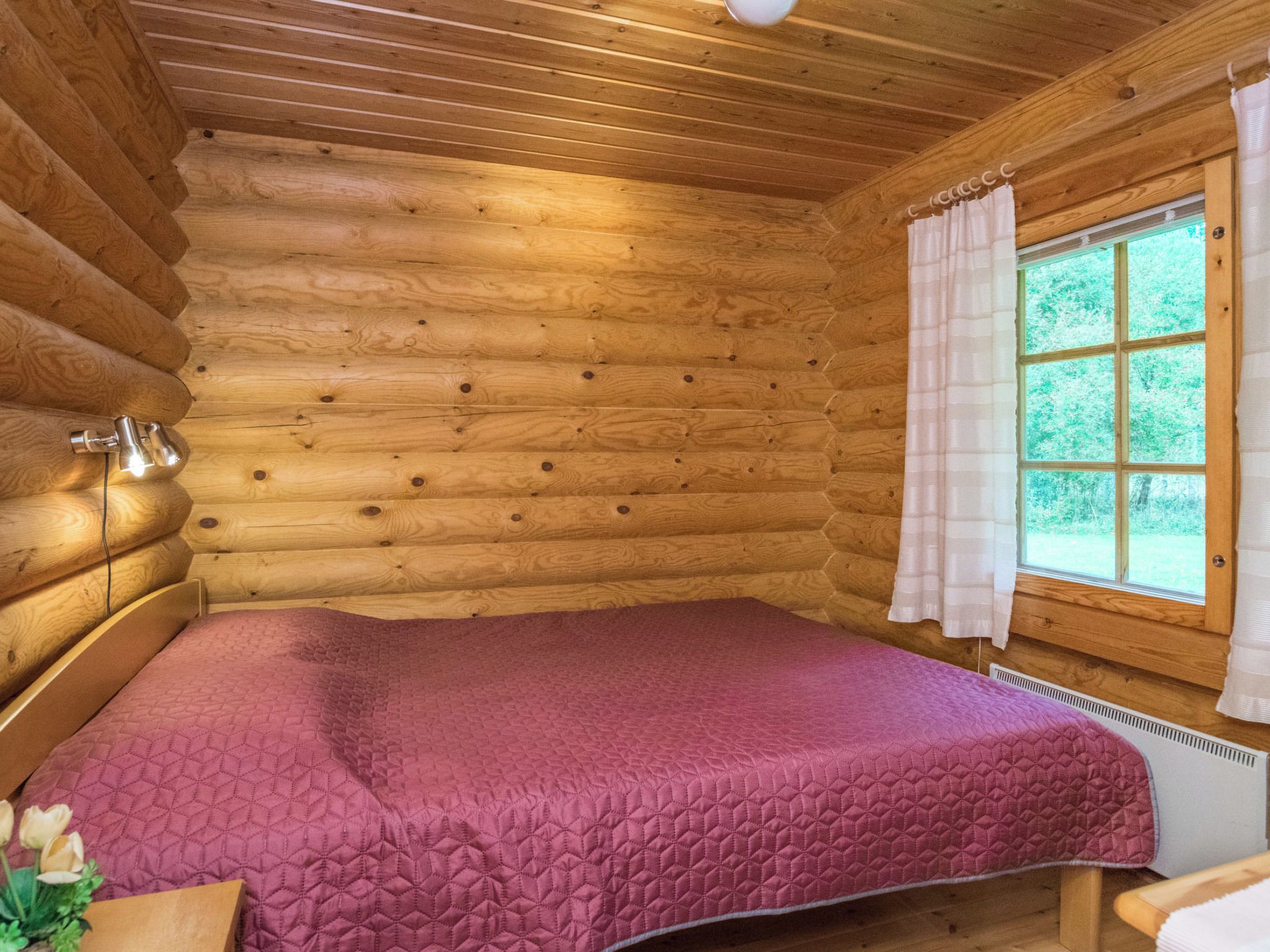 A log cabin room with a bed, a window with curtains, and a wall-mounted light fixture.