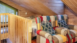 Ein gemütliches Blockhaushaus-Interieur mit einer Holztreppe, einem farbenfrohen Sessel und einem Bett mit Patchworkdecke.