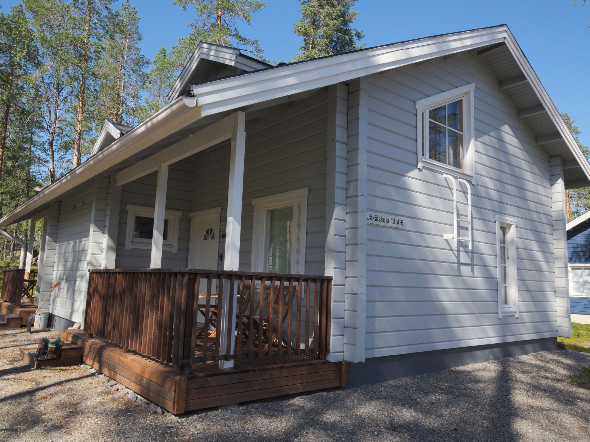 A white wooden cabin with a porch, a wooden deck, and a gravel driveway.