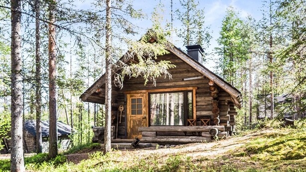 A rustic log cabin with a wooden porch and a chimney, surrounded by tall trees and greenery. - Tievatupa 2, pienempi by Interhome (Ylläsjärvi)