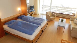 Ein Hotelzimmer mit zwei Betten, einem Schreibtisch mit Computer, einem Sessel und einer Couch, mit Blick auf ein Gebäude.