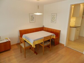Ein Essbereich mit einem Holztisch und einer Bank, einem kleinen Beistelltisch mit Lampe sowie einer Tür, die in eine Küche führt.