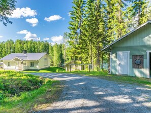 A two-story house with a porch and a garage, surrounded by a forest and a gravel driveway.