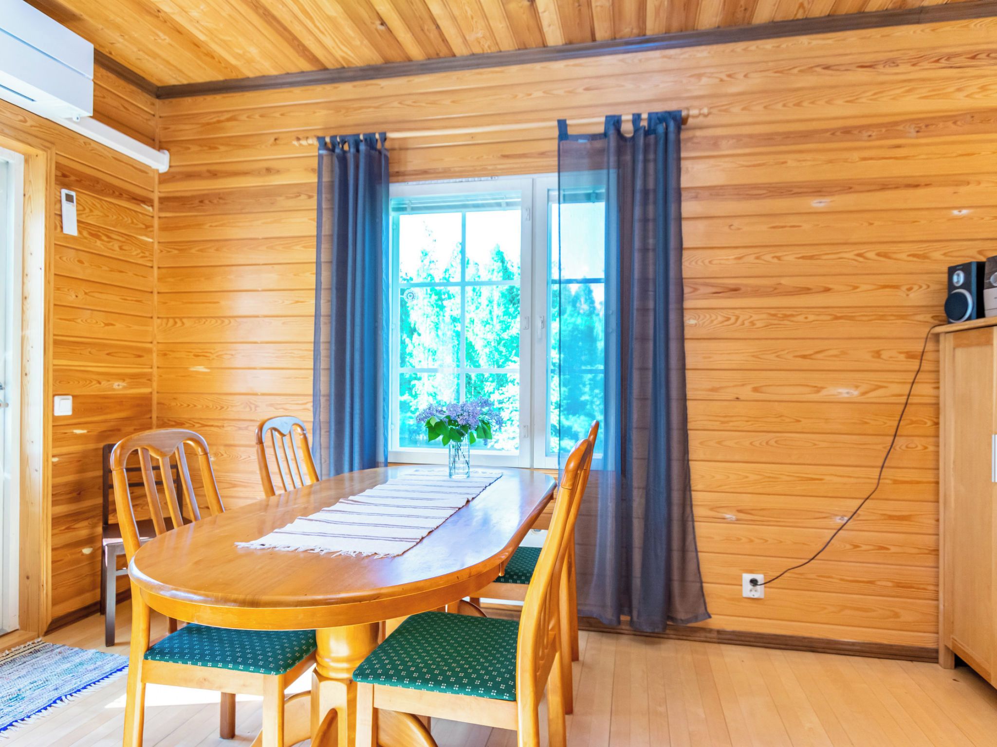 A wooden-paneled room with a dining table, chairs, a window with curtains, and a speaker on a cabinet.