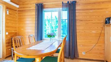 A wooden-paneled room with a dining table, chairs, a window with curtains, and a speaker on a cabinet.