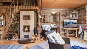 Een rustiek houten interieur met een stenen open haard, een houten trap en een gezellige zithoek met een schommelstoel en kussens.