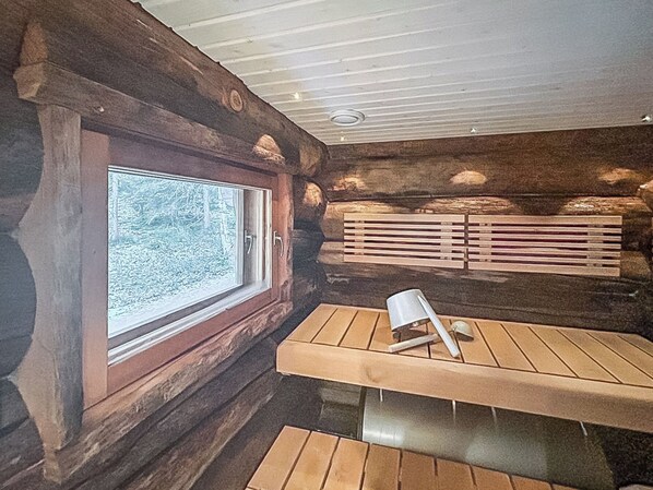 A log cabin interior with a wooden table and bench, a window with a view of trees, and a built-in bench with horizontal slats.