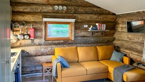 L’intérieur d’un chalet confortable, avec une cheminée, un canapé, une petite table, une fenêtre, une étagère remplie de livres et un téléviseur fixé au mur.