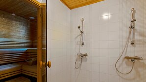 Un sauna avec des bancs en bois et une porte en verre, des murs recouverts de carreaux blancs et un espace douche équipé d’un pommeau de douche à main et d’un robinet mural.