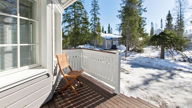 Une terrasse en bois avec une balustrade blanche, une chaise en bois et, au fond, une maison dont le toit est recouvert de neige.