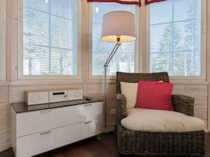 Une pièce avec un fauteuil en osier, une commode blanche, une lampe et une fenêtre avec des stores.