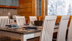 A dining area with a wooden table and white chairs, a window with a view of snow-covered trees, and a kitchen area with blue cabinets and a tiled backsplash.