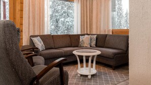 A cozy cabin interior with a wooden ceiling, a brown sectional sofa, a round coffee table, and a view of snow-covered trees through large windows.