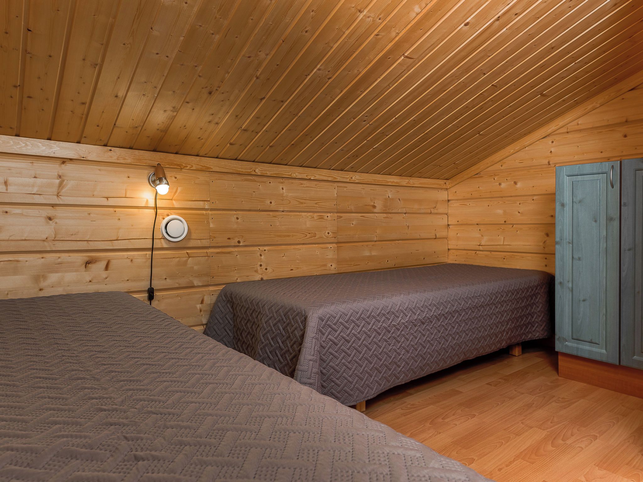 Ein kleiner, gemütlicher Raum mit Holzwänden und schräger Decke. Es gibt zwei Betten mit grauer Bettwäsche, eine Wandleuchte und einen blauen Schrank.