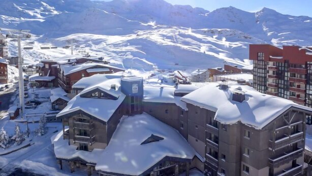 A snow-covered mountain town with buildings, including a hotel with balconies and a ski lift in the background. - Lac du Lou 506 by Interhome (Val Thorens)