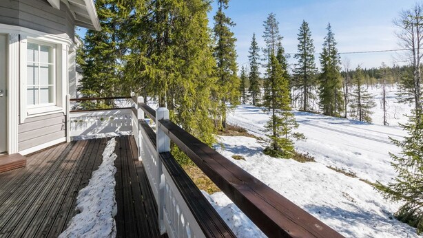 Eine Holzterrasse führt zu einem Haus, im Hintergrund eine verschneite Landschaft und immergrüne Bäume.