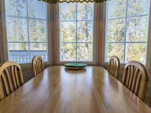 Ein Esszimmer mit einem Holztisch und -stühlen, großen Fenstern mit Jalousien und Blick auf Bäume.