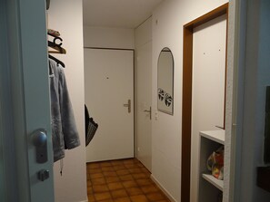 Un corridoio con una porta, un appendiabiti, uno specchio e uno scaffale con vari oggetti.