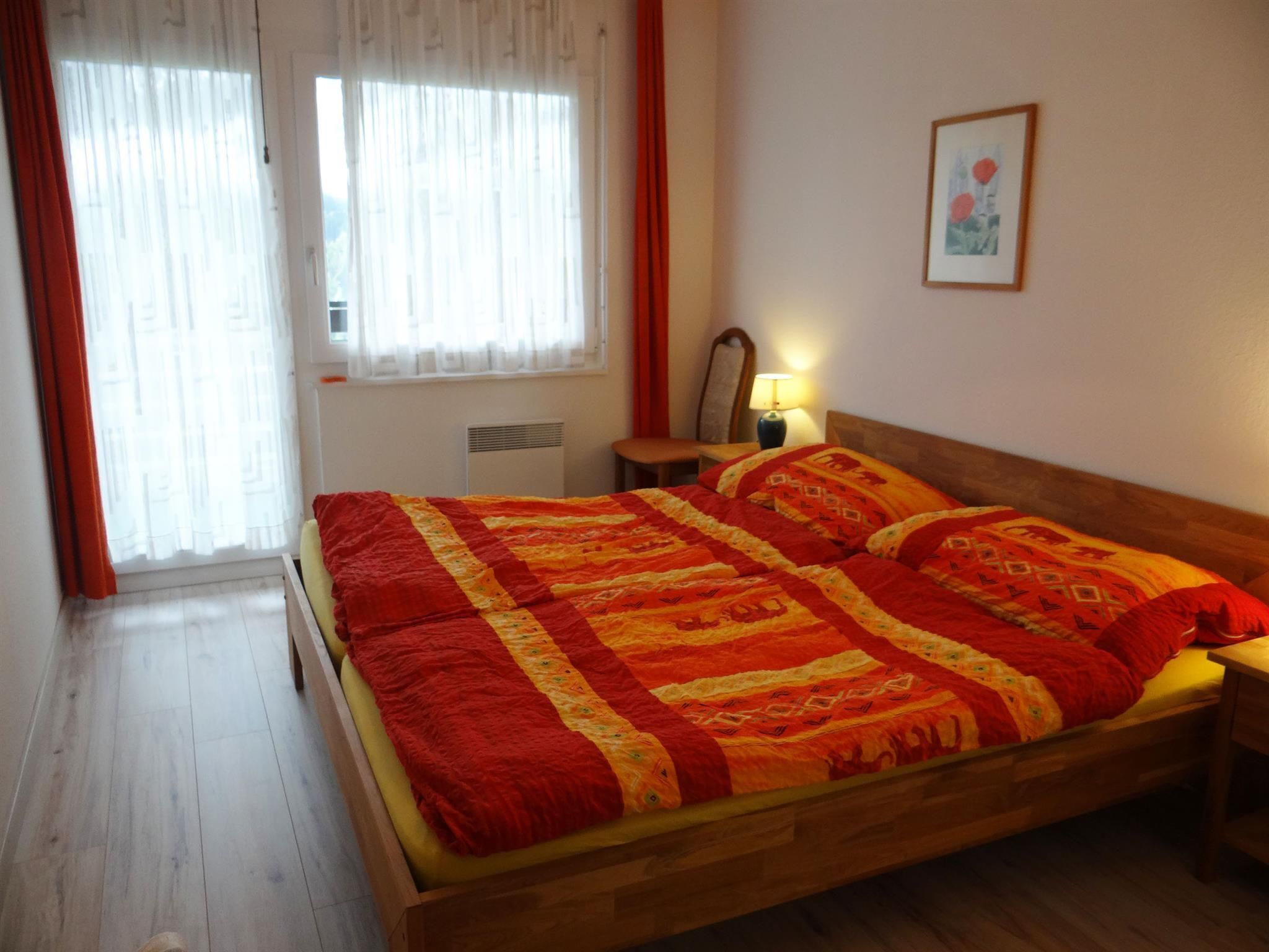 A bedroom with a wooden bed, a nightstand, a lamp, a chair, and a window with curtains.