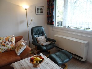 Un salon comprenant un canapé, un fauteuil, une table avec un panier de fruits, un radiateur et une fenêtre avec des rideaux.