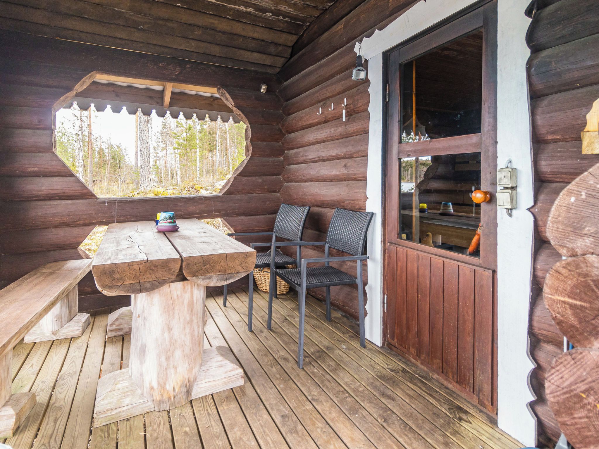 A wooden cabin with a wooden deck, a table with chairs, and a window overlooking a forest.