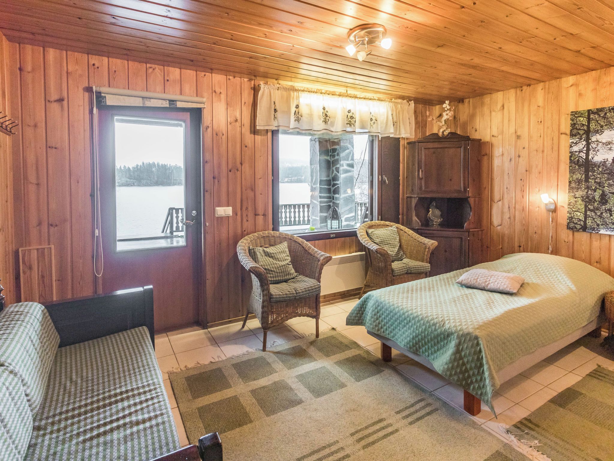 Ein Zimmer in einer Holzhütte mit einem Bett, zwei Korbstühlen, einer Bank, einem Fenster mit Vorhängen und Blick auf einen See.