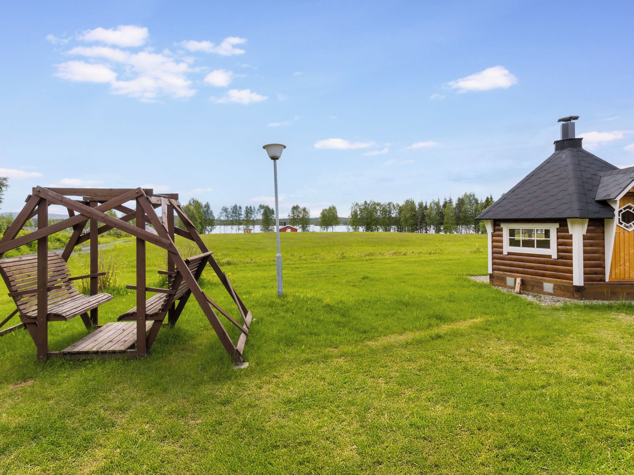 Een houten schommel, een klein huisje met schoorsteen en een lantaarnpaal in een grasveld.