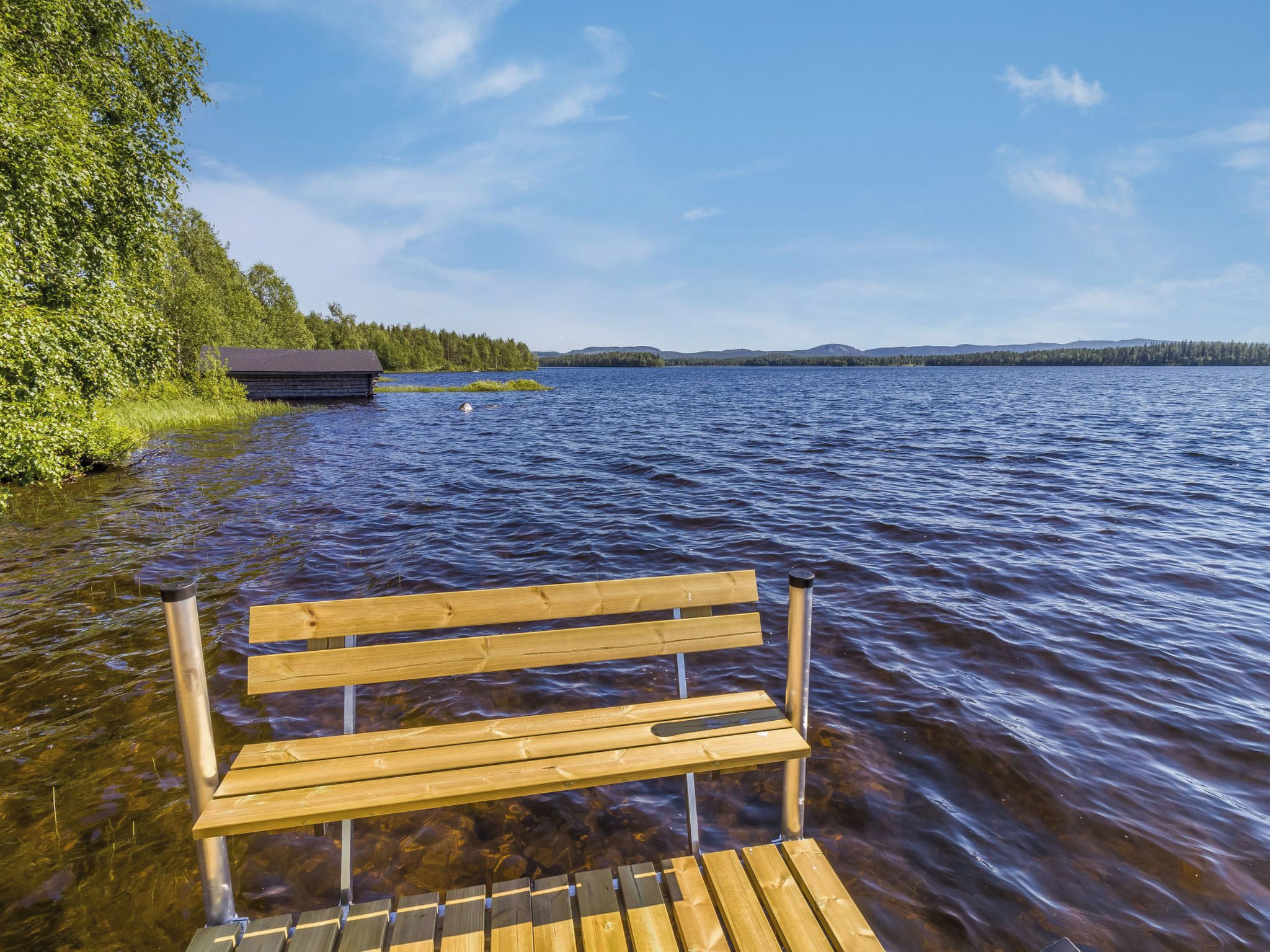 A wooden bench by a lake with a cabin in the background.