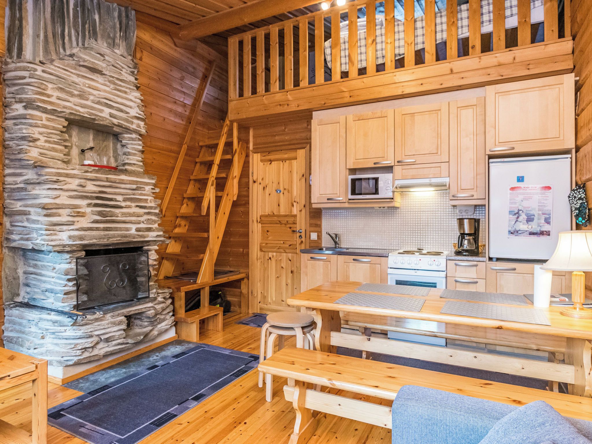 L’intérieur chaleureux d’un chalet avec une cheminée en pierre, un escalier en bois, un coin cuisine et une table à manger avec des chaises.