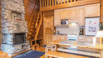 L’intérieur chaleureux d’un chalet avec une cheminée en pierre, un escalier en bois, un coin cuisine et une table à manger avec des chaises.