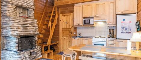 A cozy cabin interior with a stone fireplace, wooden staircase, kitchen area, and a dining table with chairs.