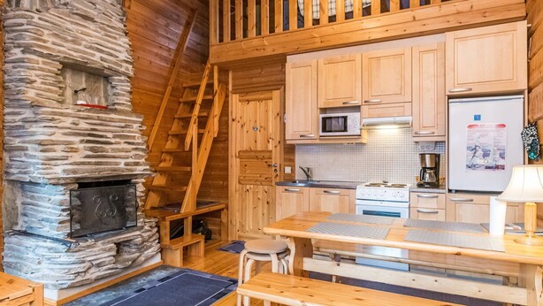 A cozy cabin interior with a stone fireplace, wooden staircase, kitchen area, and a dining table with benches. - Rukansyli 1 by Interhome (Kuusamo)