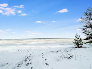 Schneebedeckte Landschaft mit Fußspuren, die zu einer Baumgrenze und einem Gewässer führen.