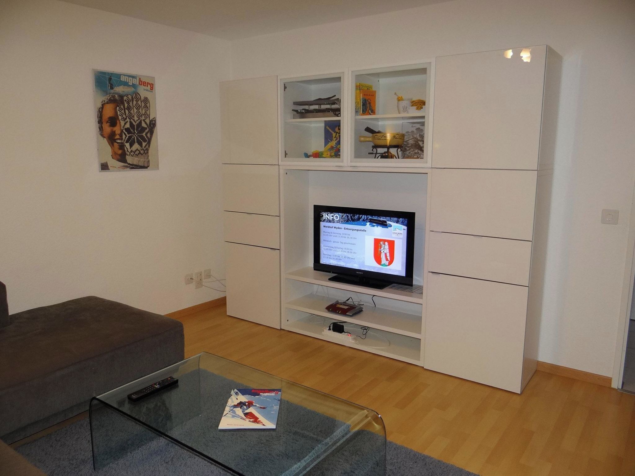 Ein Wohnzimmer mit einem Flachbildfernseher auf einem Standfuß, einem Glastisch mit einer Zeitschrift, einem grauen Sofa und einer weißen Schrankwand mit verschiedenen Gegenständen.