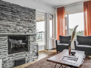 A modern living room with a stone fireplace, two grey armchairs, a wooden coffee table, and a view of the outdoors through large windows.